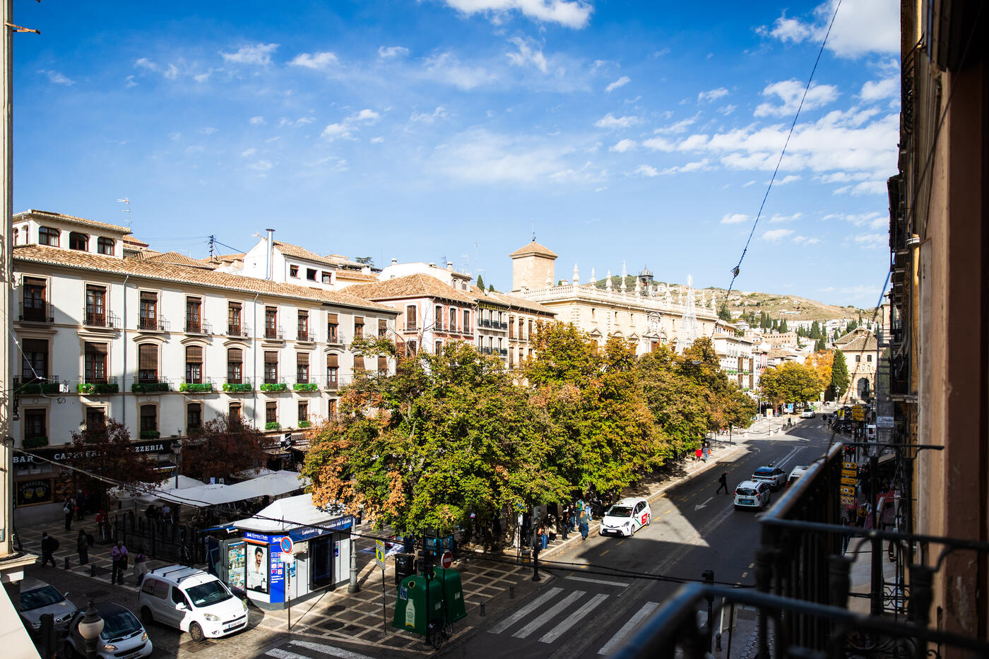 Plaza Nueva a mediodía desde el balcón de la Suite Comares: la fila de bares con sus toldos blancos, los plátanos de sombra, el quiosco de la plaza y, al fondo, la Real Chancillería y la ladera del Albaicín.